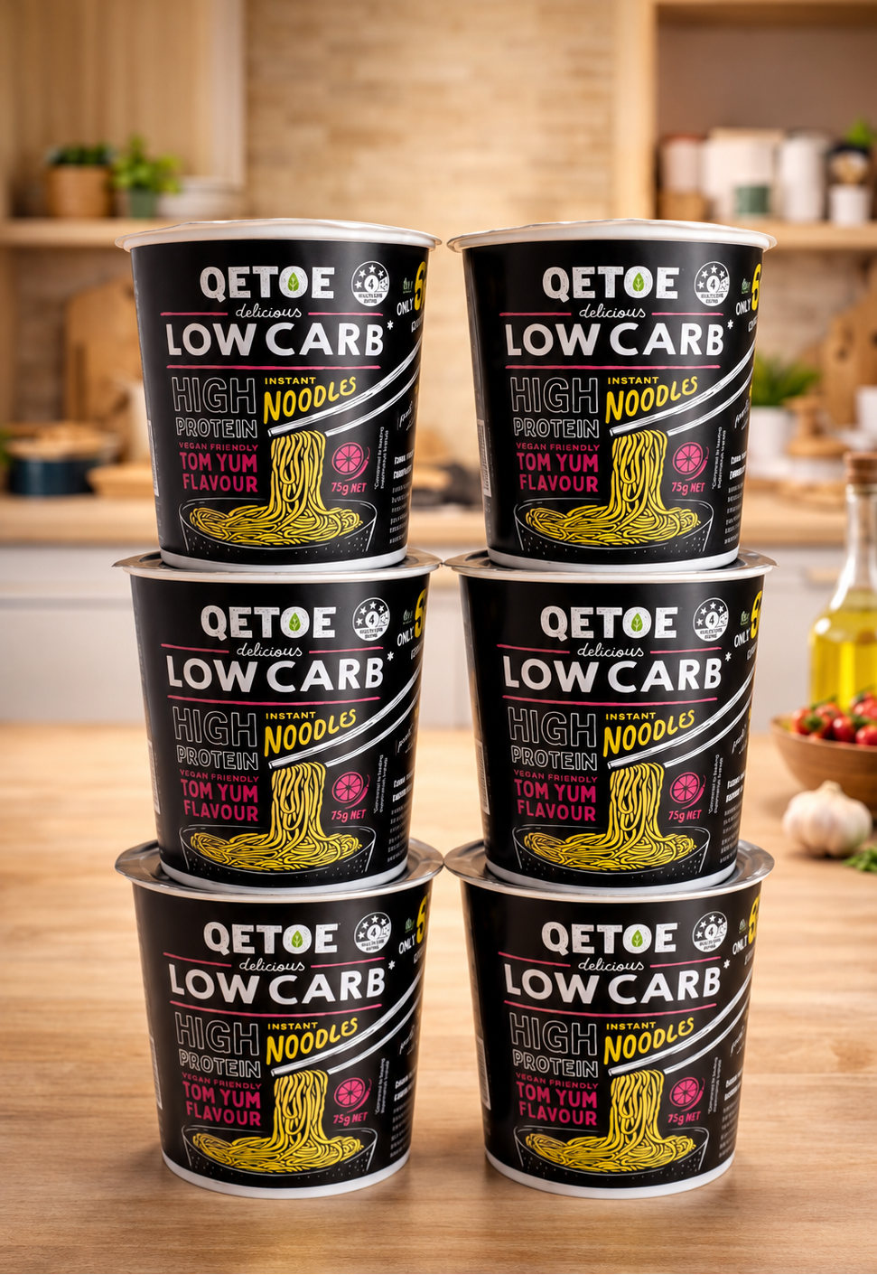 Six QTOE Low Carb High Protein Noodles cups stacked in two columns of three on a wooden kitchen counter, with a blurred kitchen background.