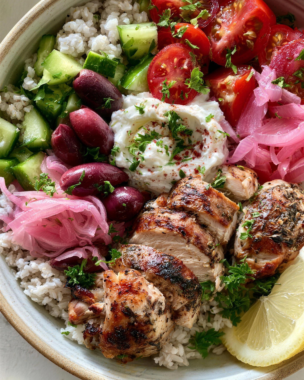 A bowl of Greek chicken over white rice with tomatoes, cucumber, olives, pickled onion and whipped feta